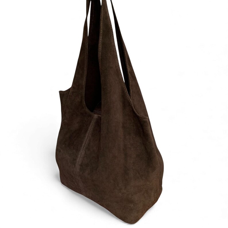K&ouml;nnte beinhalten: Eine dunkelbraune Hobo-Tasche aus Wildleder mit einer lockeren, unstrukturierten Silhouette. Die Tasche verf&uuml;gt &uuml;ber einen breiten, in den K&ouml;rper integrierten Schulterriemen, sichtbare vertikale Nahtdetails und eine weiche, matte Textur.