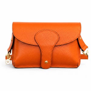 May include: A bright orange leather handbag with a pebbled texture, featuring a fold-over flap with a central tab closure secured by a gold-tone metal stud. The bag includes gold-tone hardware attachments on the sides and a small circular charm with a W.
