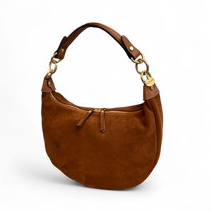 May include: A brown suede shoulder bag with a curved design. It has a gold zip, gold chain details, and a brown leather handle. The bag features a single compartment and a small, round, gold-toned emblem.