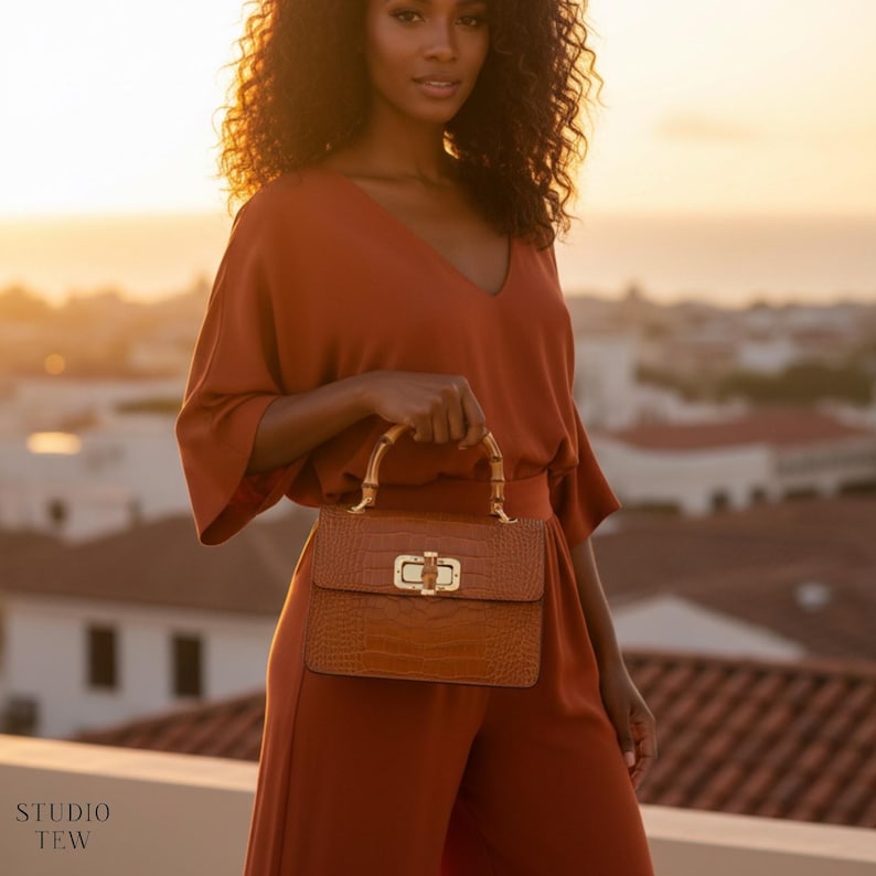 Puede incluir: Un bolso de mano marr&oacute;n estructurado con un acabado texturizado de imitaci&oacute;n cocodrilo, con un cierre de metal dorado y un asa de bamb&uacute; curvada. El bolso se sostiene frente a un conjunto de color &oacute;xido s&oacute;lido sobre un fondo desenfocado.