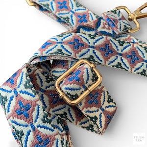 May include: A woven bag strap featuring a repeating geometric pattern in shades of dusty rose, deep teal, and bright blue on a cream background. The strap includes gold-toned metal hardware, including a rectangular adjustable slider and a swivel hook.