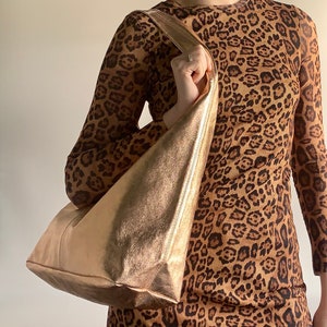 May include: A gold-coloured leather tote bag with a triangular shape. The bag is being held by a person wearing a leopard print dress.