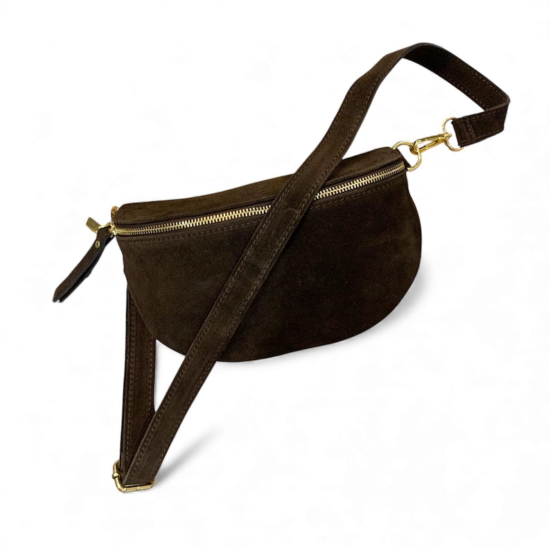 May include: A dark brown suede fanny pack with a gold zipper and hardware. The adjustable strap is also dark brown. The bag is curved and has a minimalist design, suitable for carrying essentials.