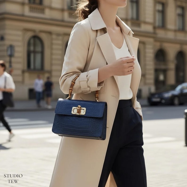 Puede incluir: Un bolso de mano azul marino estructurado con un acabado texturizado de piel de cocodrilo, que presenta un cierre giratorio de metal dorado y dos asas de estilo bamb&uacute;. El bolso se lleva con una gabardina beige, top blanco y pantal&oacute;n negro.