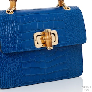 Puede incluir: Un bolso de mano azul vibrante con un patr&oacute;n texturizado de relieve de cocodrilo. El bolso incluye un cierre de metal dorado con un pasador central estilo bamb&uacute; y un asa de bamb&uacute; a juego. El texto STUDIO TEW es visible en la esquina inferior.