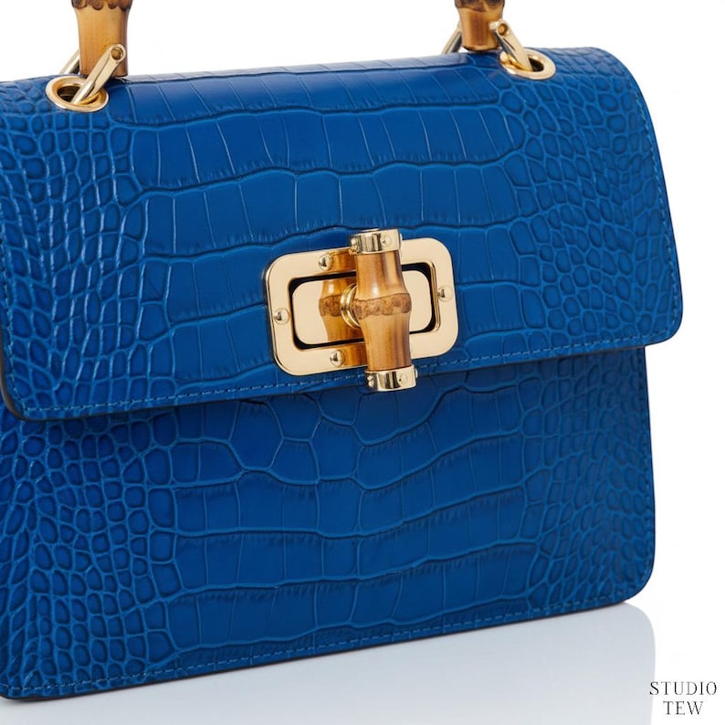 Puede incluir: Un bolso de mano azul vibrante con un patr&oacute;n texturizado de relieve de cocodrilo. El bolso incluye un cierre de metal dorado con un pasador central estilo bamb&uacute; y un asa de bamb&uacute; a juego. El texto STUDIO TEW es visible en la esquina inferior.