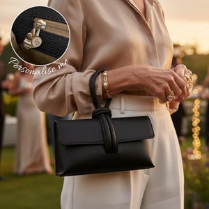 May include: A black textured leather handbag with a single top handle and a flap closure. An inset circular detail shows a silver zipper pull with a white circular charm featuring the letter S. The text "Personalise me" is written in white script.