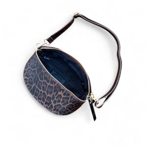 Puede incluir: Un bolso bandolera en forma de media luna con un estampado de leopardo en tonos marr&oacute;n y negro. El bolso cuenta con un cierre de cremallera dorado, un forro interior oscuro y una correa marr&oacute;n ajustable sujeta con clips met&aacute;licos dorados.