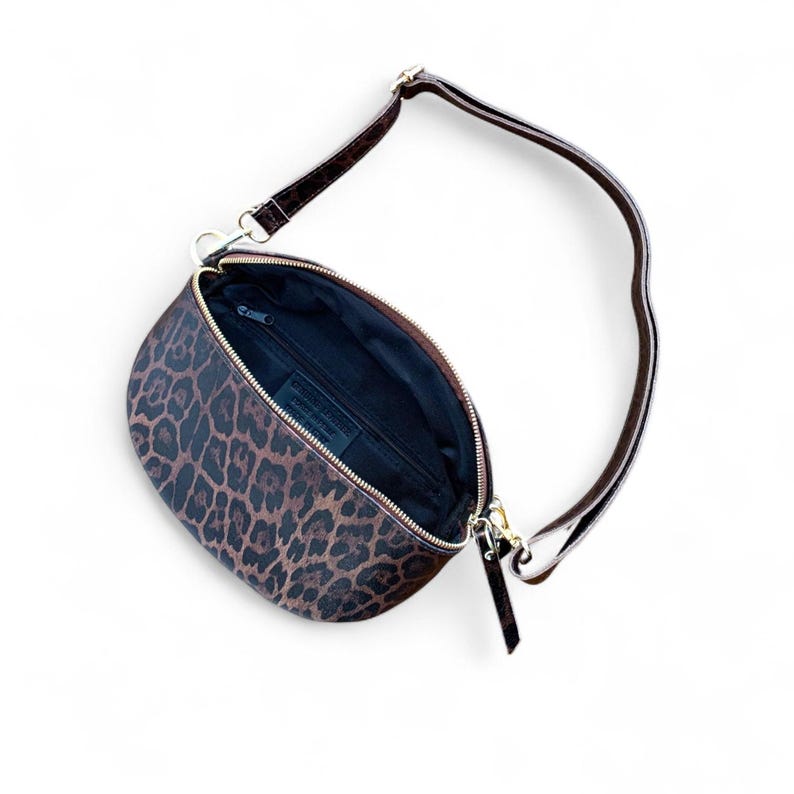 Puede incluir: Un bolso bandolera en forma de media luna con un estampado de leopardo en tonos marr&oacute;n y negro. El bolso cuenta con un cierre de cremallera dorado, un forro interior oscuro y una correa marr&oacute;n ajustable sujeta con clips met&aacute;licos dorados.