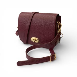 Puede incluir: Bolso bandolera de cuero burdeos con acabado texturizado. El bolso presenta un cierre y herrajes dorados, una correa larga ajustable y un asa más corta. El bolso tiene un cierre de solapa y una forma redondeada.