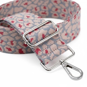 May include: A woven bag strap featuring a repeating animal print pattern in shades of dusty pink, grey, and vibrant red. The strap includes a rectangular silver-toned metal slider for length adjustment and a silver-toned metal lobster clasp hook.