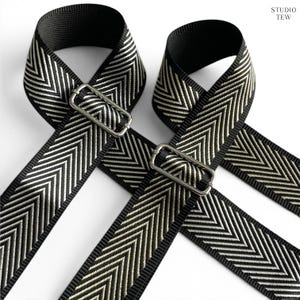 Peut inclure: Deux bandoulières réglables avec un motif chevron noir et argent métallisé. Chaque sangle comprend une boucle coulissante en métal argenté pour le réglage. Le texte STUDIO TEW est imprimé dans le coin supérieur droit de l'image.