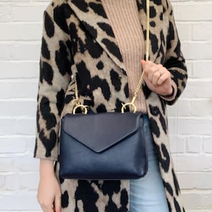 May include: A navy blue leather satchel bag with a gold chain strap. The bag has a flap closure and is worn over the shoulder of a person wearing a leopard print coat.