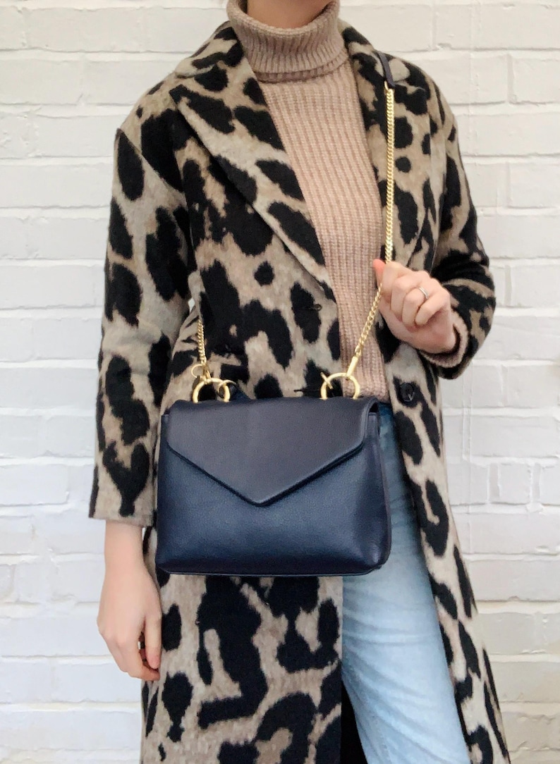 May include: A navy blue leather satchel bag with a gold chain strap. The bag has a flap closure and is worn over the shoulder of a person wearing a leopard print coat.