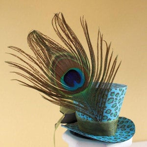 May include: A miniature top hat with a teal leopard print design, adorned with a large peacock feather. The hat features a green ribbon band and brim, creating a whimsical and decorative accessory.