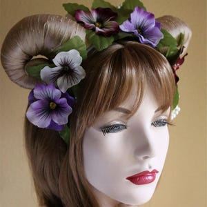 May include: A decorative headpiece featuring curved, horn-like structures wrapped in a tan fabric. The headpiece is adorned with purple and white pansy flowers and green leaves, creating a whimsical and fantasy-inspired look.