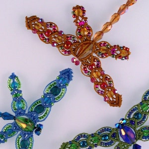 Op de afbeelding: Drie kralenlibellen-sieraden in blauw, groen en oranje met iriserende accenten. De libellen zijn gemaakt van kleine kralen en draad.