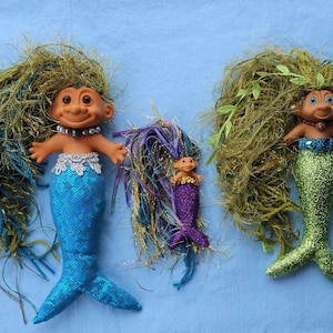 May include: Three mermaid troll dolls with long, colorful hair. The largest doll has blue hair and a turquoise tail, the middle doll has purple hair and a purple tail, and the smallest doll has green hair and a green tail.