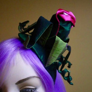 May include: A decorative headpiece featuring a large, dark green bow with a contrasting pink and green flower accent. The headpiece is set against a background of purple hair and a neutral backdrop, creating a striking visual contrast.