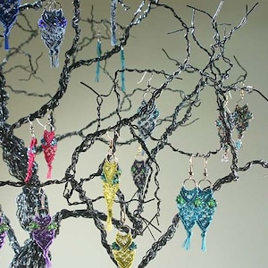 Op de afbeelding: Een display van handgemaakte macramé oorbellen in diverse kleuren, waaronder blauw, groen, roze en oranje, hangend aan een decoratieve draadboom. De oorbellen hebben ingewikkelde knopen en kralenwerk.