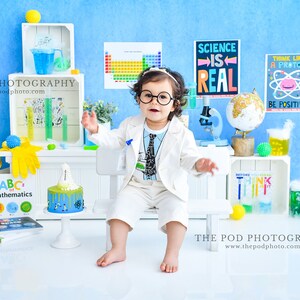 May include: A young child dressed as a scientist wearing a white lab coat and a blue and white tie, sitting on a white stool in front of a blue background. The child is holding a beaker and a test tube. There are various science-themed decorations around the child, including a periodic table, a globe, and a microscope. The text "SCIENCE IS REAL" is visible on a poster behind the child.