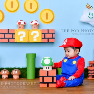 May include: A baby dressed in a red and blue Mario costume sits on a wooden floor. The baby is looking at a green mushroom toy. The background is a blue wall with a brick wall, green pipes, and gold coins. The text 