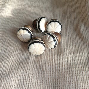 May include: Six decorative cabinet knobs. Each knob has a cream-coloured, flower-like design with black stripes and a brown base. The knobs are arranged in a circular pattern on a textured, beige fabric.