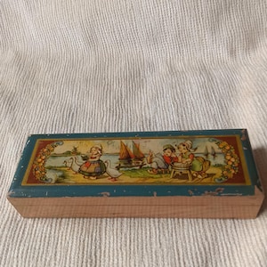 Peut inclure: Une boîte rectangulaire en bois avec un couvercle à charnière présentant une illustration colorée d'enfants en vêtements traditionnels près d'un plan d'eau avec des voiliers et des oies. La boîte a une bordure peinte bleu sarcelle usée.