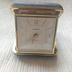 Puede incluir: Una taza de cerámica blanca con un asa negra y el texto "But First, Coffee" en negrita negra. La taza se encuentra sobre un fondo borroso, sugiriendo un ambiente acogedor.