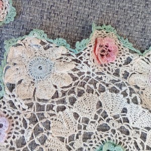 May include: Close-up of handmade crochet doilies. The doilies feature a floral design with pastel pink, blue, and green accents. The edges are scalloped, and the intricate openwork creates a delicate, vintage aesthetic.
