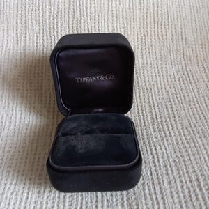 May include: A black velvet jewellery box with a hinged lid. The interior features a black velvet ring slot and a satin-lined lid with the text Tiffany & Co. printed in silver lettering. The exterior of the box has a smooth, dark finish.
