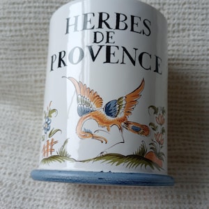 May include: A white ceramic cylindrical canister with a blue base rim. The front features the black text "HERBES DE PROVENCE" above a hand-painted illustration of an orange and blue bird in flight, surrounded by small green and blue floral motifs.