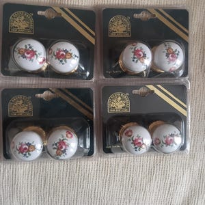 May include: Four sets of two round white ceramic cabinet knobs with gold-tone bases, each set in individual plastic packaging. The knobs feature a floral pattern with red, pink, and yellow roses. The packaging is labeled Grove Brand Hardware.