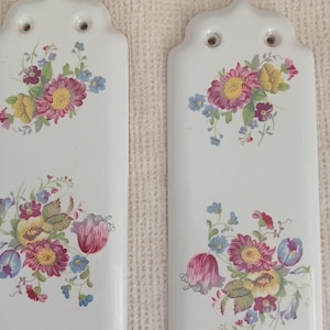 以下が含まれることがあります： 白いセラミック製の壁掛けプレート2枚。花柄のデザインです。各プレートには、ピンク、黄色、青の花束が描かれています。プレートの上部には装飾があり、吊り下げ用の穴が2つあります。