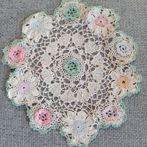 May include: A handmade, off-white crochet doily with a floral design. The doily features small, colorful crochet flowers in shades of pink, blue, and yellow, with a green scalloped edge. The doily is placed on a gray surface.