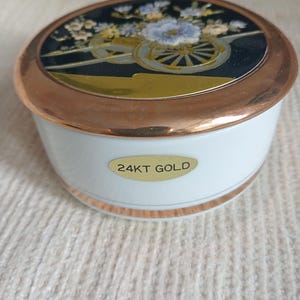 Puede incluir: Caja de baratijas de cerámica blanca con borde y tapa dorados. La tapa presenta un diseño floral con fondo negro y dorado. La caja tiene una etiqueta dorada que dice "24KT GOLD".