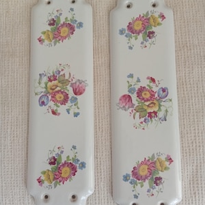 Op de afbeelding: Twee witte keramische deurvingerplaten met bloemmotieven. Elke plaat toont een boeket van roze, gele en blauwe bloemen. De platen hebben een decoratieve bovenkant en drie gaten voor montage. Een vintage-stijl woondecoratie-item.