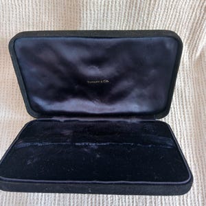 May include: An open, rectangular, black velvet jewelry box with a navy blue satin interior. The box has a hinged lid and the words "Tiffany & Co." are printed in gold lettering inside the lid.