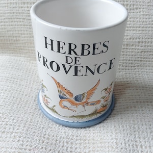 May include: A white ceramic cylindrical container with a blue base rim. The front features the black text "HERBES DE PROVENCE" above a colorful illustration of a bird in flight surrounded by small floral accents and green foliage details.