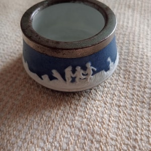 May include: A small, blue ceramic bowl with a white relief design of figures. The bowl has a dark rim and a light blue interior. The design wraps around the base of the bowl. The bowl is sitting on a textured surface.