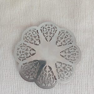 May include: A silver-colored, decorative coaster with an intricate, openwork floral design. The coaster has a scalloped edge and a polished, reflective surface. The design features a symmetrical pattern of swirling leaves and vines.