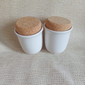 Può includere: Due contenitori in ceramica bianca con coperchi in sughero. I contenitori hanno una forma arrotondata e una finitura liscia e lucida. I coperchi in sughero sono circolari e si adattano perfettamente. Lo sfondo è un tessuto strutturato bianco sporco.