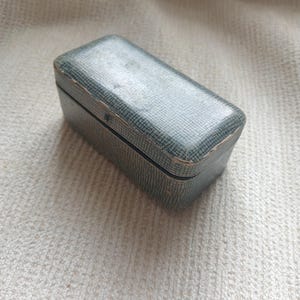 Puede incluir: Una caja rectangular vintage con un exterior texturizado de color verde azulado. La caja tiene una tapa con bisagras y parece estar hecha de un material similar al cuero. La caja está sobre una superficie texturizada de color neutro.