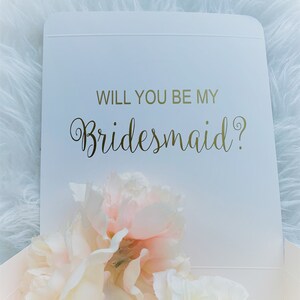 May include: White box with a gold inscription that reads "Will you be my bridesmaid?"  The box is open and contains a bouquet of white and pink flowers.