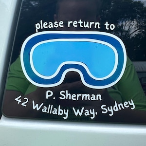 May include: A white sticker on a car window features a blue and white design resembling goggles. The text "please return to" is above the goggles, with "P. Sherman" and address "42 Wallaby Way, Sydney" below.