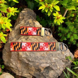 May include: Two keychains with a Maryland flag design. The larger keychain is red, black, orange, and white with a gold metal ring. The smaller keychain is the same design but smaller. Both keychains are on a brown rock.