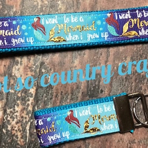 Puede incluir: Dos llaveros azules y morados con un diseño de sirena y el texto "Quiero ser una sirena cuando crezca". Los llaveros tienen anillas de plata.
