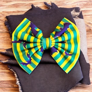 May include: A large, decorative bow with a Halloween theme. The bow features a pattern of yellow and green stripes with purple bat silhouettes. The bow is centered on a dark grey fabric with frayed edges, set against a wooden background.