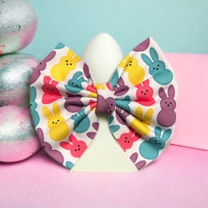 May include: A white bow with a pastel pink, yellow, purple, and blue bunny pattern. The bow is tied around a white egg-shaped object.