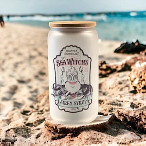 May include: A white frosted glass jar with a wooden lid. The jar has a label that says "Atlantica Apothecary The Sea Witch's Siren Syrup 1836". The label features a cartoon image of Ursula, the sea witch from Disney's The Little Mermaid.